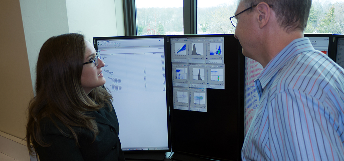 Two medical professionals stand close together in front of several screens, discussing the information displayed.