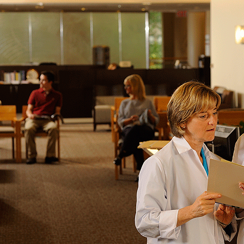 A photograph of the inside of a building with two people sitting in chairs, and a doctor in the forefront reading a file.