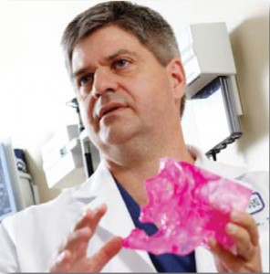 A photograph of a doctor speaking with his hands and showing a pink device to someone off camera.