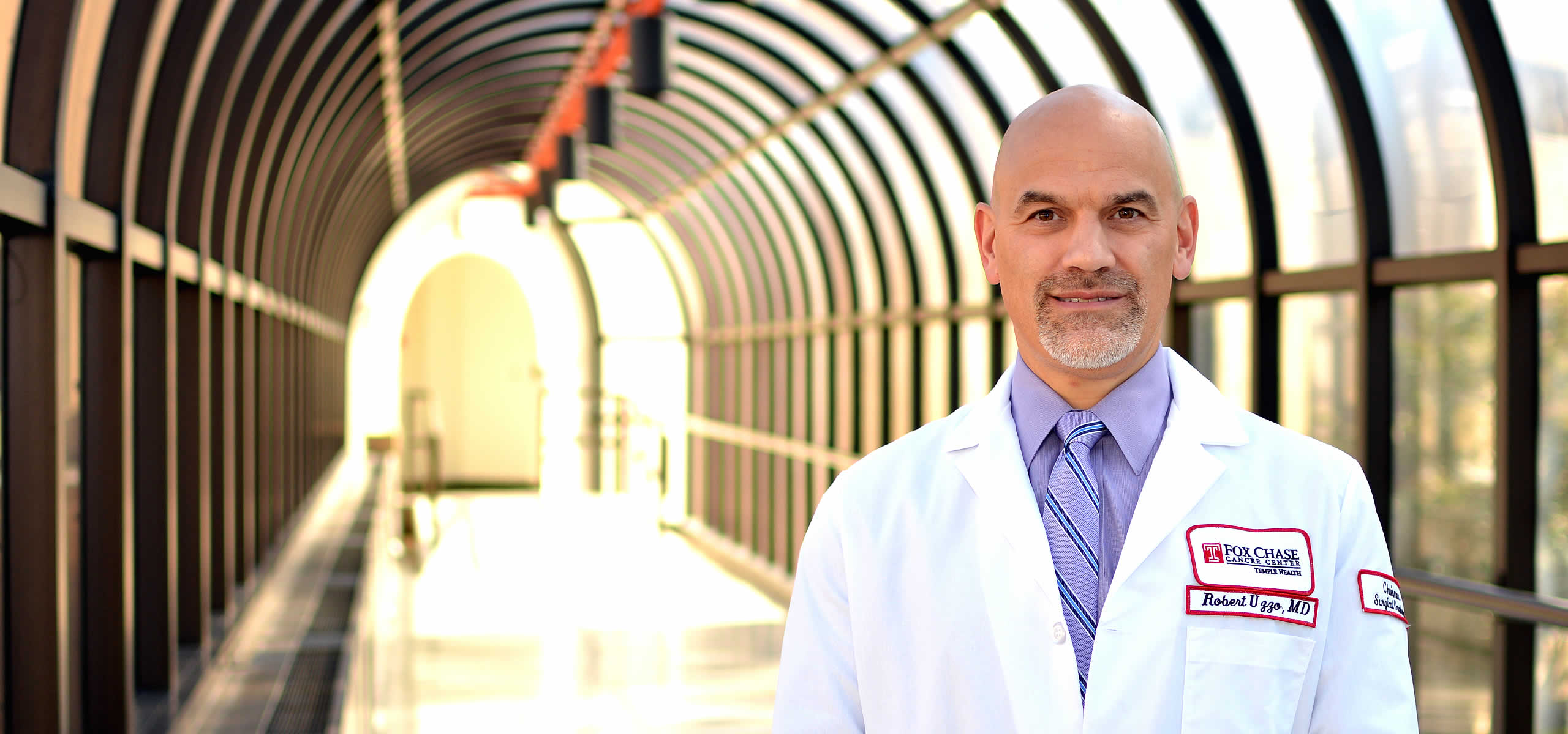 A portrait shot of Dr. Robert Uzzo, standing on the right side of the frame in a blurry hallway with lots of windows.