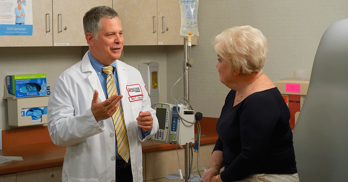 A photograph of a Fox Chase doctor smiling as he speaks with a patient receiving an IV in a medical room.