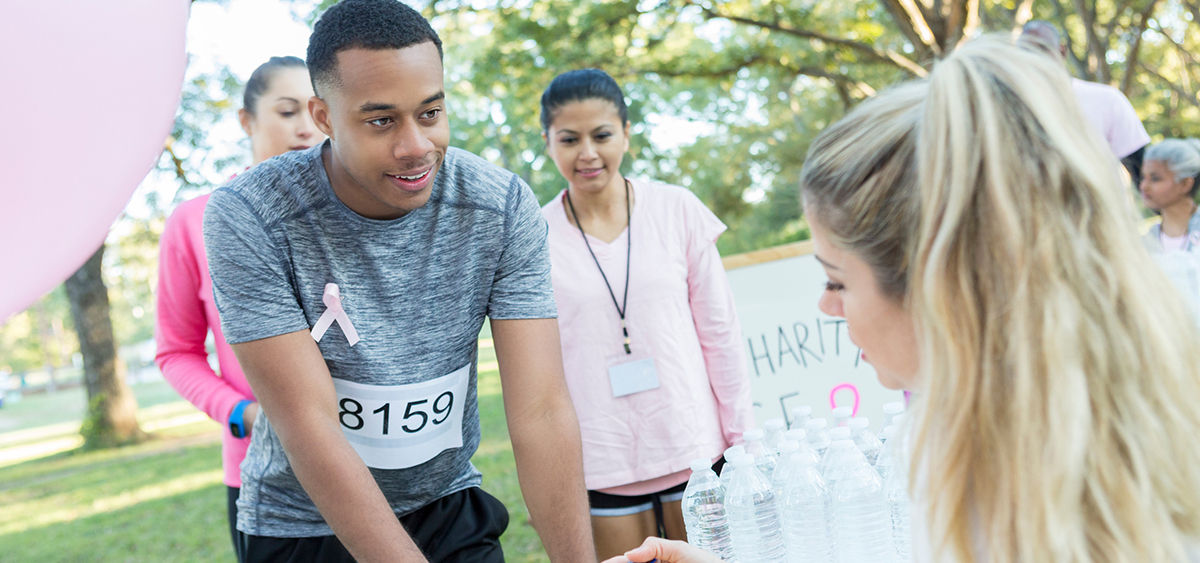 A photograph of three runners at a table with water bottles on it, with a person behind the table speaking to one of them.