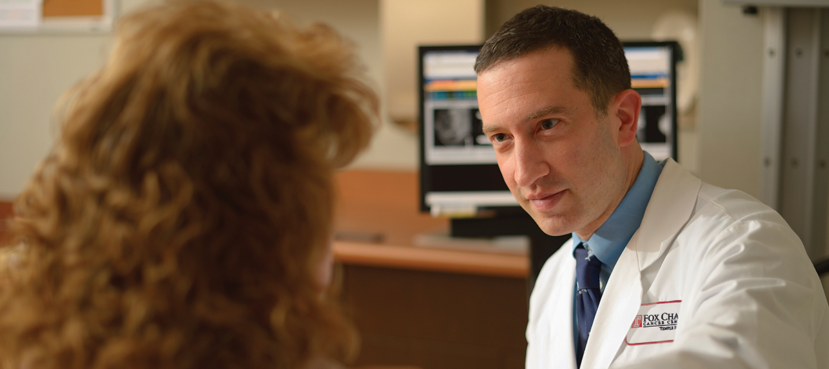 A closeup photo looking over a person's shoulder, as a Fox Chase doctor leans close while they talk in a medical room.