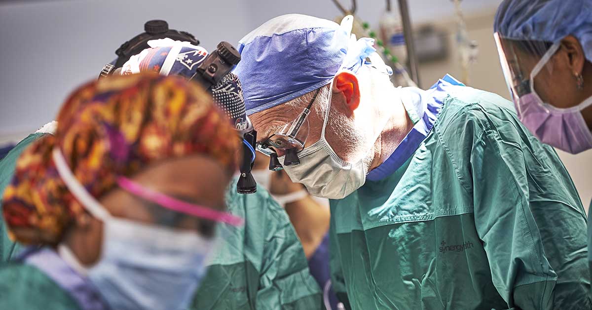 A group of doctors in full surgical gear all look down at the same thing, what they're doing obscured from view.