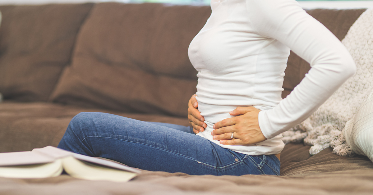 A photograph of a person sitting on a couch in jeans and a white long sleeve, clutching at their lower side with both hands.