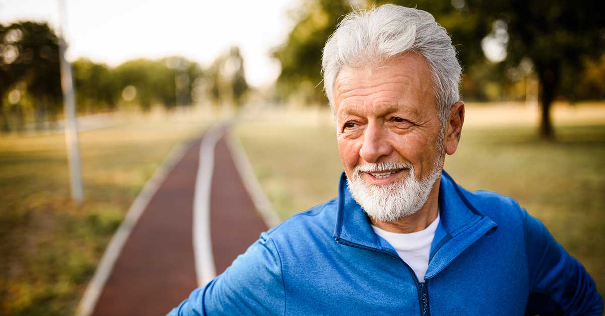 A portrait shot of a man with his hands on his hips standing on a running track, smiling as he looks off camera.