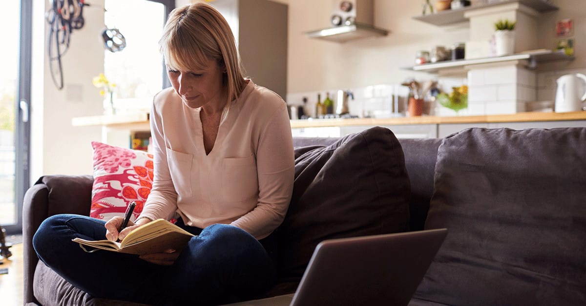 A photo of a woman sitting on the couch with a laptop next to her, holding a pen and pad of paper in her lap.