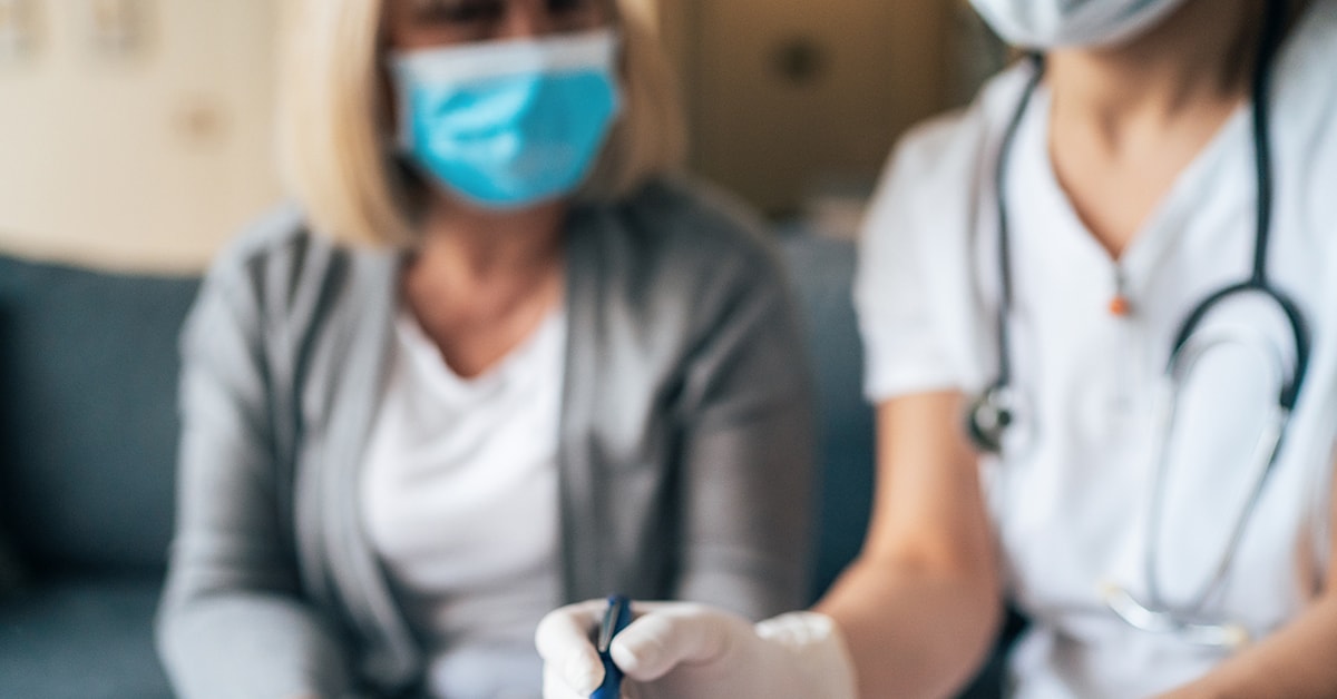 A blurred photo of a medical professional and a woman both wearing masks as they sit together and work on something.