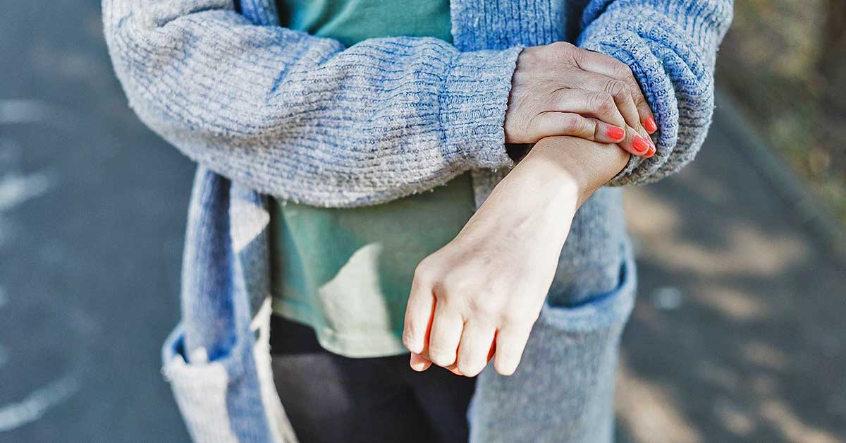 A closeup photo of a person's extended arm, which they touch with their other hand as they stand outside.