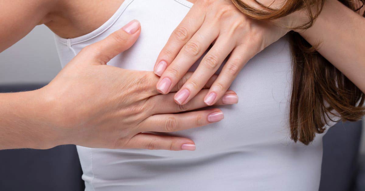 A close of up a person wearing a white tank top, with both of their hands over one their breasts.