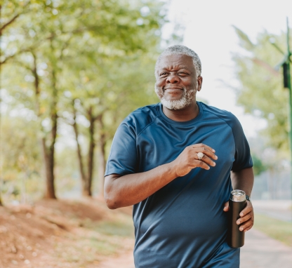 An older Black man smiling while jogging on a tree-lined path, holding a reusable water bottle.