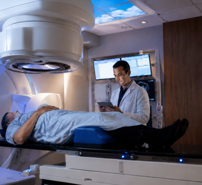 A patient lies on a treatment table beneath a radiation therapy machine while a healthcare provider reviews information on a tablet in a modern treatment room.