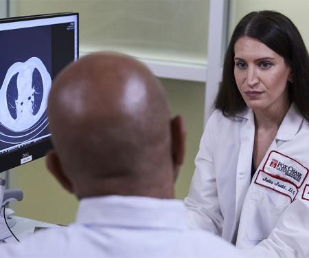 A Fox Chase doctor converses with another person in front of a screen with results from a medical imaging test on it.