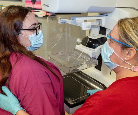 A medical professional and a patient stand in front of a mammogram machine, wearing masks and looking at each other.