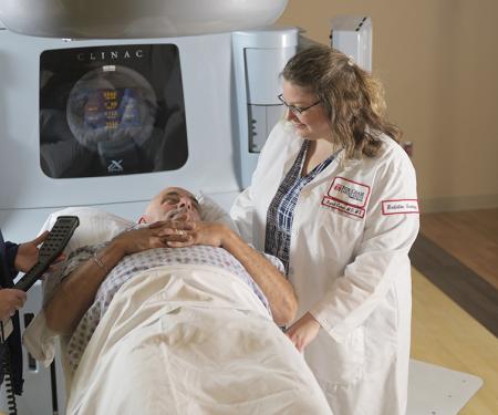 A photo of two Fox Chase medical professionals smiling at patient laying in a bed in front of a large radiation therapy machine.