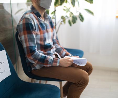 A photograph of a person sitting in a teal chair in the corner of a room, holding a paper and looking off camera.