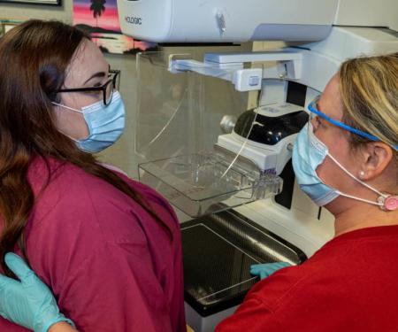 A medical professional and a patient stand in front of a mammogram machine, wearing masks and looking at each other.