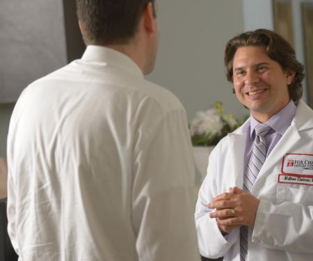 A photograph of a Fox Chase doctor smiling widely at someone, with another person on the phone at a desk to their left.