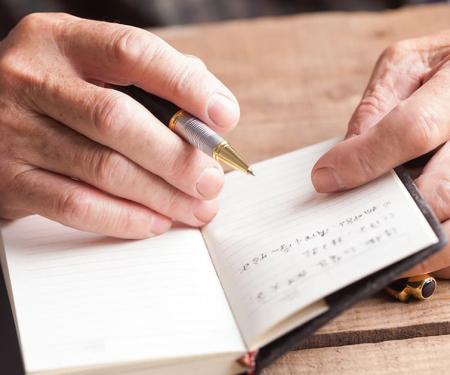A closeup photo of a person's hands, holding a pen as they write in a small notebook.
