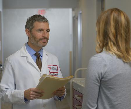 A Fox Chase doctor stands facing another person in a hallway, holding a file in his hands.