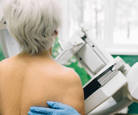 A photograph from behind a person receiving a mammogram, with a gloved hand on their back.