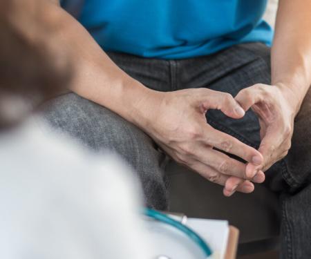 A closeup photo looking over a doctor's shoulder to a person's lap, where they loosely intertwine their fingers together.