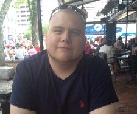 A photograph of  Andrew Steinmetz sitting at an outdoor table, with a phone in hand and a drink in front of him.