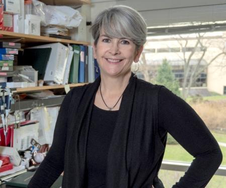 A photograph of Denise Connolly standing in a cluttered laboratory, smiling at the camera.