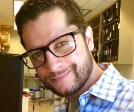 A closeup photo of Janusz Franco-Barraza smiling at the camera, standing in a room full of files and equipment.