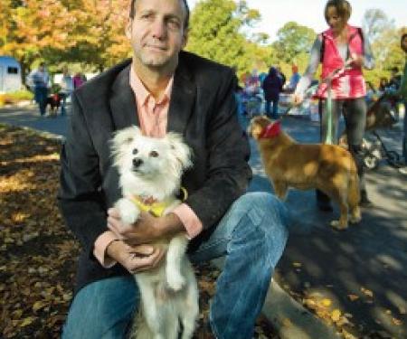 A photograph of Frank Serianni outside with several other people in the background, kneeling down and holding his white dog.