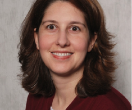 A portrait shot of a person with shoulder length brown hair wearing a dark red shirt, smiling at the camera.