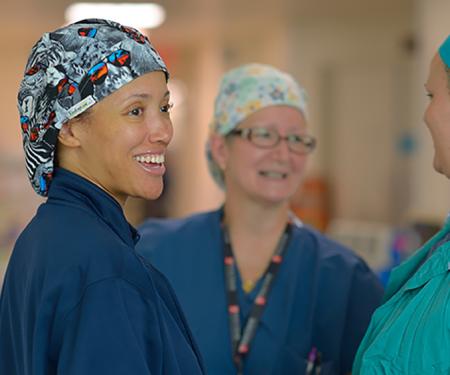 A photograph of three medical professionals wearing hair coverings smiling as they speak to each other in a hospital hallway.