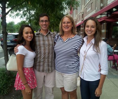 A photograph of a family of four smiling at the camera on a street lined with shops, a mother, father, and two teenage daughters