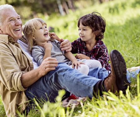 A photograph of an older man sitting in a grassy field with two children on his lap, all smiling widely.