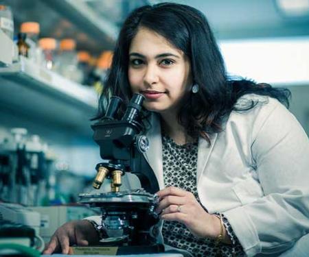A photograph of Dr. Sanjeevani Arora, PhD, holding a microscope close to her and looking up at the camera.