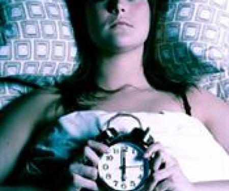 A photograph of a person laying in bed in a dark room, looking up at the ceiling with an alarm clock in their hands.