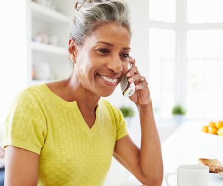 A photo of a person standing in their kitchen with a phone up to their ear and a laptop in front of them.