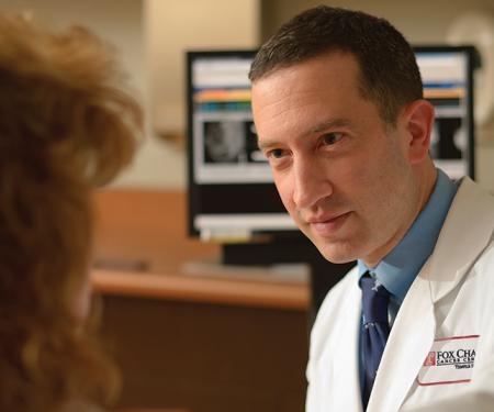 A closeup photo looking over a person's shoulder, as a Fox Chase doctor leans close while they talk in a medical room.