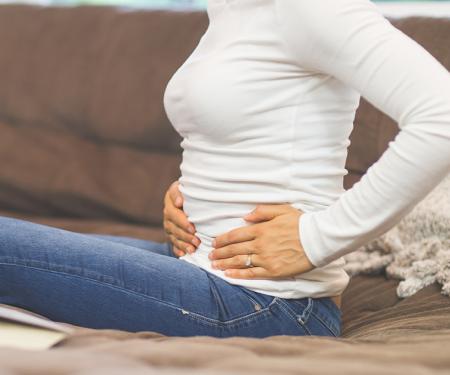 A photograph of a person sitting on a couch in jeans and a white long sleeve, clutching at their lower side with both hands.