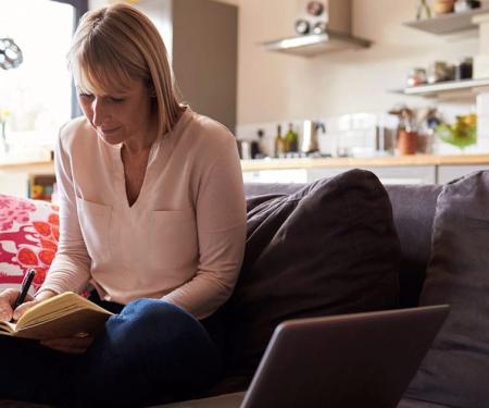 A photo of a woman sitting on the couch with a laptop next to her, holding a pen and pad of paper in her lap.