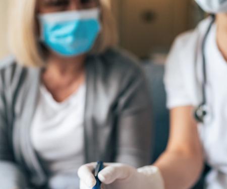 A blurred photo of a medical professional and a woman both wearing masks as they sit together and work on something.