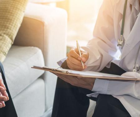 A photograph of a medical professional sitting across from a patient, taking notes on a clipboard.