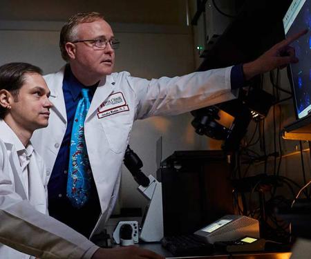 A photograph of two Fox Chase doctors sitting in a dim room, one gesturing at a screen with various blue splotches displayed.