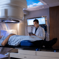 A patient lies on a treatment table beneath a radiation therapy machine while a healthcare provider reviews information on a tablet in a modern treatment room.