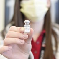 A photograph with a blurred background of a person holding a vial that reads &quot;COVID-19 Vaccine&quot;.