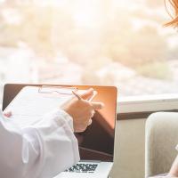 A photo from behind two people, with a clipboard and laptop in front of them and bright sunlight coming in from the window.