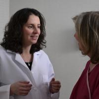A photograph of a medical professional standing close to a patient in a medical chair, as they smile and speak with each other.