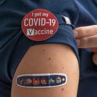 A close-up photograph of a bandaid on a person's upper arm, with a sticker that says &quot;I got my COVID-19 vaccine&quot; on their shirt.