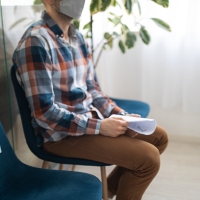 A photograph of a person sitting in a teal chair in the corner of a room, holding a paper and looking off camera.
