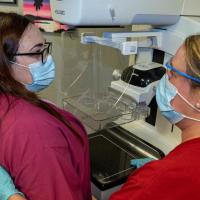 A medical professional and a patient stand in front of a mammogram machine, wearing masks and looking at each other.